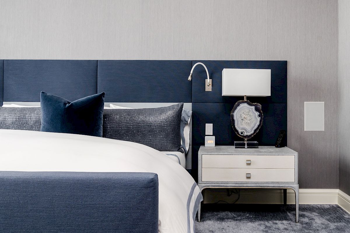 A modern bedroom with a blue headboard, white bedding, nightstand, lamp, and decor against a gray wall.