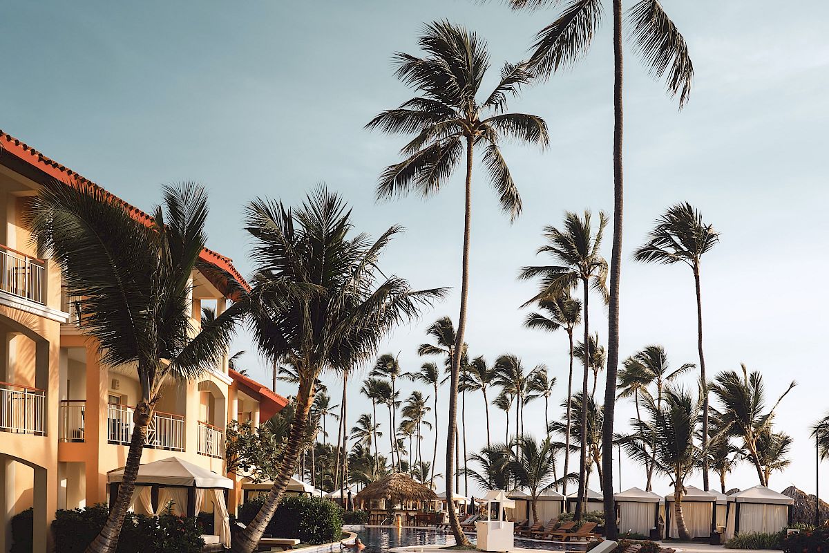 The image shows a poolside area with lounge chairs, palm trees, and a building under a blue sky, creating a tropical resort vibe.