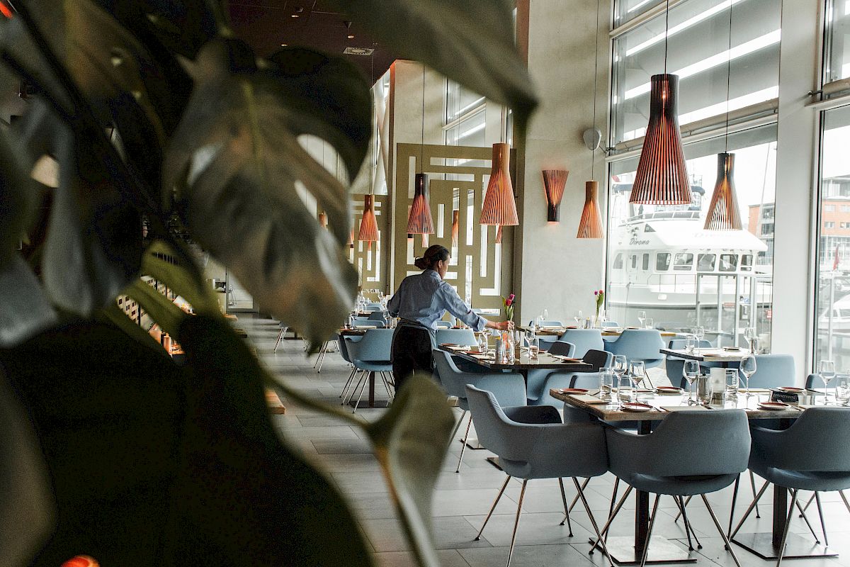 A modern restaurant interior with a person setting tables, stylish lighting, and large windows dominate the scene.