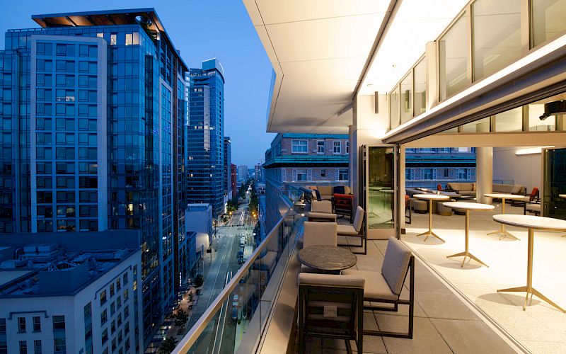 A modern cityscape at dusk, featuring a rooftop terrace with seating and tables, overlooking tall buildings and a lit street below.